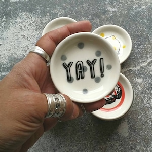 May include: A set of four small ceramic dishes with different designs. One dish has a white background with black polka dots and the word "YAY!" written in black. Another dish has a white background with a red and black design. The third dish has a white background with a yellow and black design. The fourth dish has a white background with a black and white design.
