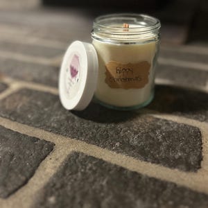 May include: A white candle in a glass jar with a brown label that says "hippy christmas". The candle is sitting on a brick surface.