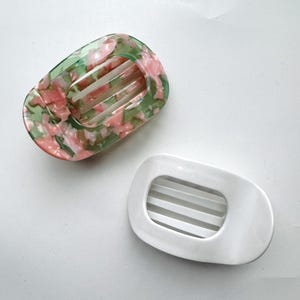 May include: Two soap dishes. One is a marbled green and pink floral pattern. The other is solid white. Both have an oval shape with a rectangular opening and horizontal bars for drainage. The dishes are designed to hold soap.