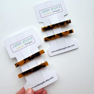 May include: Two rectangular hair barrettes with a tortoiseshell pattern are displayed on white cards. The cards have the text "candy argyle designs" and the website address. The barrettes are brown and black.