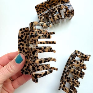 May include: Three leopard print hair claw clips in varying sizes. The clips are made of a glossy, tortoiseshell-like material with black and tan spots. The clips are open, showing the teeth that hold the hair in place. The background is white.