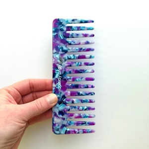 May include: A wide-toothed comb with a marbled pattern in shades of blue, purple, and white. The comb is held by a hand against a white background. The brand name "candy argyle" is printed on the comb.