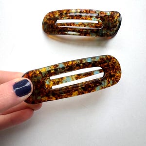 May include: Two rectangular hair clips with rounded edges, featuring a translucent tortoiseshell pattern in shades of brown, amber, and light blue. Each clip has a central horizontal cutout, creating a frame-like design with a smooth, glossy finish.