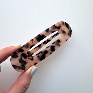 May include: Oval-shaped hair clip with a tortoiseshell pattern. The clip features a light brown base with dark brown spots. A long, narrow opening runs down the center of the clip.