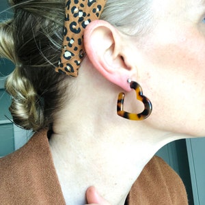 May include: Tortoiseshell heart-shaped earrings. The earrings are a warm brown color with dark brown spots. A brown and black leopard print headband is also visible.