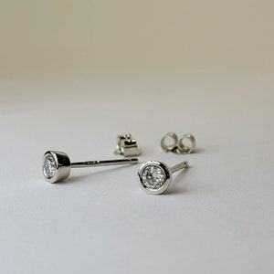 Op de afbeelding: Een paar zilveren oorstekers met een bezel-zetting, elk met een heldere, ronde edelsteen. De oorbellen worden met hun achterkanten getoond, wat een minimalistisch ontwerp laat zien. De zetting is helder, glanzend zilver.