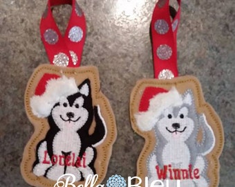 May include: Two felt Christmas ornaments shaped like dogs wearing Santa hats. The ornament on the left has a black and white dog with the name "Lonelai" embroidered on it. The ornament on the right has a gray and white dog with the name "Winnie" embroidered on it.