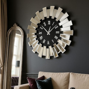 Może przedstawiać: Duży, okrągły zegar ścienny z czarną tarczą i srebrnymi rzymskimi cyframi. Zegar otoczony jest prostokątami z luster, tworząc efekt promieni słonecznych. Zegar wisi na ciemnoszarej ścianie nad beżową sofą.