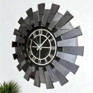May include: A contemporary wall clock featuring a black and silver sunburst design. The clock face is black with white Roman numerals and hands. The clock is mounted on a white wall, adding a modern touch to any room.