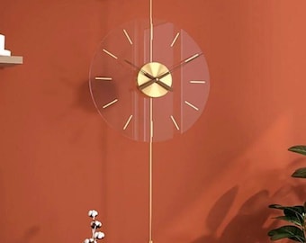 May include: A modern wall clock with a transparent face and gold-colored hands and accents. The clock hangs from a wooden mount and features a long, decorative pendulum. The clock is set against a burnt orange wall.