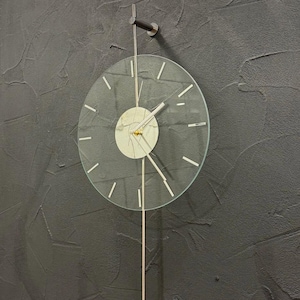 Può includere: Un orologio da parete moderno con quadrante in vetro trasparente e design minimalista. L'orologio presenta una cornice e lancette color argento, con un lungo e sottile pendolo che termina con un accento rosso. Montato su una parete strutturata grigio scuro.