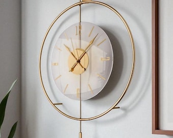 May include: A gold-colored, circular wall clock with a minimalist design. The clock features a clear glass face with gold hands and hour markers. A gold pendulum hangs below the clock face. The clock is mounted on a light gray wall.