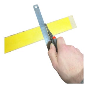 Pu&ograve; includere: Una mano usa un taglierino per tagliare una striscia gialla di materiale, guidata da un righello di metallo. Il righello &egrave; contrassegnato da misure in centimetri. Lo sfondo &egrave; bianco.