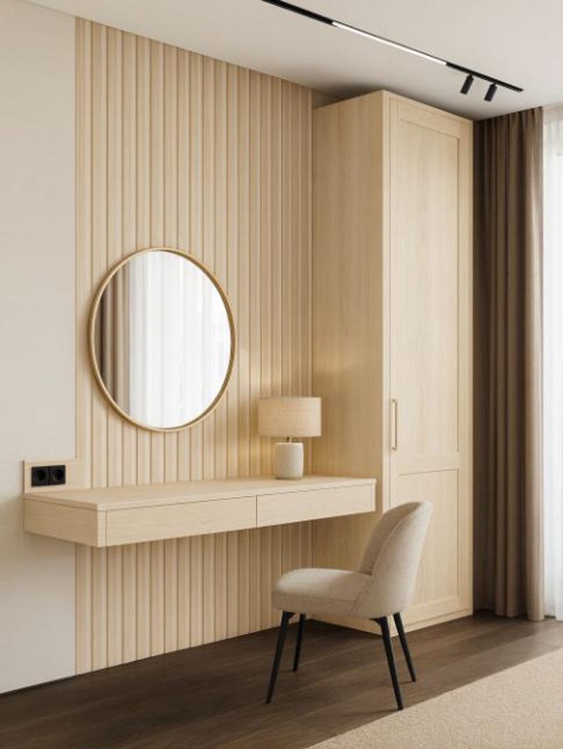 May include: A modern bedroom interior featuring a light wood vanity with a round mirror and a small lamp. A beige upholstered chair with black legs sits in front of the vanity. The wall has vertical wood paneling, and a tall cabinet is visible.
