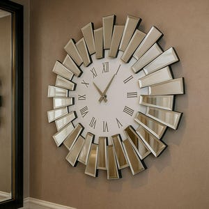 Może przedstawiać: Dekoracyjny zegar ścienny z białą tarczą i czarnymi rzymskimi cyframi. Zegar otoczony jest prostokątnymi lustrzanymi akcentami, tworzącymi wzór promieni słonecznych. Zegar jest zamontowany na beżowej ścianie.