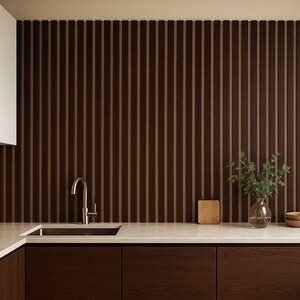 May include: Modern kitchen design featuring dark brown wood cabinetry and a matching vertical slat wall. A stainless steel faucet and sink are set into a light-colored countertop. Decorative elements include a small wooden cutting board, a glass vase with greenery, and two wooden bowls.