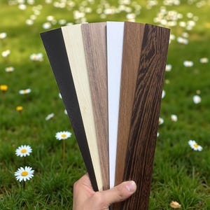 Pu&ograve; includere: Una mano tiene in mano un ventaglio di campioni di finiture effetto legno in vari colori: nero, beige chiaro, venatura del legno media, bianco, venatura del legno scura e una venatura del legno pi&ugrave; scura. Lo sfondo &egrave; un campo verde sfocato con fiori bianchi.
