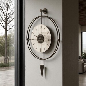 Può includere: Un orologio da parete moderno con un design circolare. L'orologio presenta un quadrante bianco con numeri e lancette nere, racchiuso in una cornice di metallo scuro. Un pendolo decorativo è appeso sotto. È montato su una parete grigio chiaro.