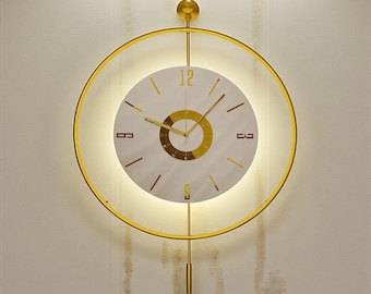 May include: A modern wall clock with a gold-colored circular frame and a white face. The clock features gold-colored hands and hour markers. The clock has a pendulum and is illuminated with a soft light.