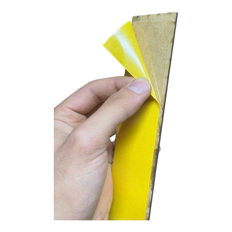 May include: A hand peeling yellow adhesive tape from a rectangular wooden board. The tape is bright yellow with a white backing, contrasting with the brown wood. The image is set against a white background.