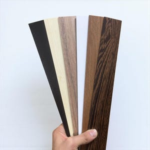 May include: A hand holding a fan of wood veneer samples in various colors and wood grain patterns. The samples include black, light beige, medium brown, white, dark brown, and a dark brown with a prominent wood grain.