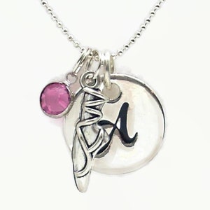 May include: Silver necklace with a charm of a ballet shoe, a small pink crystal, and a silver disc with the letter 'A' engraved on it.
