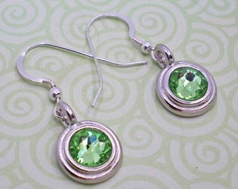 Peut inclure: Boucles d'oreilles argentées avec une pierre verte claire ronde sertie dans un cadre. Les boucles d'oreilles ont un design à crochet et sont présentées sur un fond vert clair avec un motif tourbillonnant.