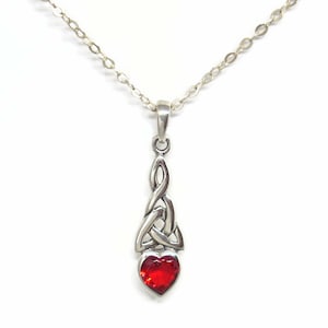 May include: A silver-toned pendant necklace featuring a Celtic knot design with a heart-shaped, red gemstone. The pendant hangs from a delicate chain against a white background. The necklace is a piece of jewelry.