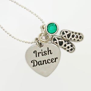 May include: Silver necklace with a heart-shaped pendant that reads "Irish Dancer." The necklace also features a green gemstone and two small shoe charms, likely representing Irish dance shoes. The chain is delicate.