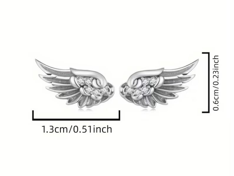 Może przedstawiać: Para kolczyk&oacute;w w kształcie skrzydeł w kolorze srebrnym z osadzonymi przezroczystymi kamieniami. Kolczyki mają 1,3 cm szerokości i 0,6 cm wysokości, co wskazują czarne linie pomiarowe i etykiety tekstowe widoczne na obrazie.