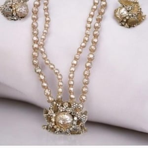 Puede incluir: Un conjunto de joyas vintage con un collar de perlas de dos hilos y pendientes a juego. El collar tiene cierres dorados ornamentados y un gran colgante central con detalles intrincados. Los pendientes tienen un diseño similar. El conjunto se muestra sobre una superficie blanca.