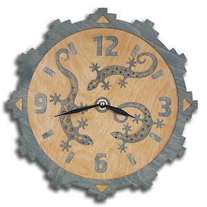 May include: A round wooden clock with a gray outer ring and a light wood face. The clock features two stylized geckos with intricate patterns and black hands. The numbers 12, 9, 6, and 3 are visible on the clock face.