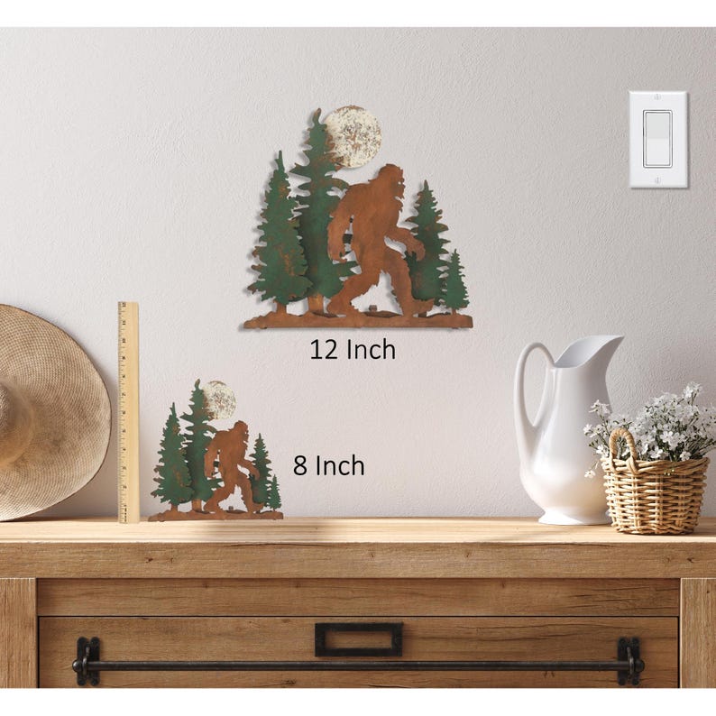 May include: Two metal wall sculptures of Bigfoot walking through a forest with a full moon. The larger sculpture is 12 inches tall and the smaller sculpture is 8 inches tall.