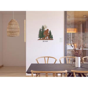 May include: Metal wall art featuring a Bigfoot silhouette walking through a forest with a full moon in the background. The art is painted in shades of brown, green, and white. The art measures 16 inches in width.