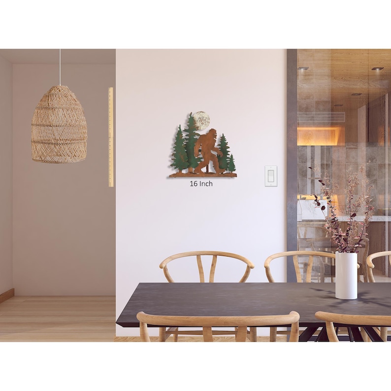 May include: Metal wall art featuring a Bigfoot silhouette walking through a forest with a full moon in the background. The art is painted in shades of brown, green, and white. The art measures 16 inches in width.