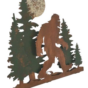 May include: Metal wall art featuring a silhouette of Bigfoot walking through a forest under a full moon. The art is painted in shades of brown, green, and white.