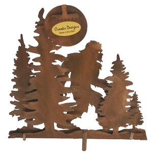 May include: Rustic metal wall decor featuring a silhouette of Bigfoot walking through a forest. The piece is made by Crooks Designs and is made in the USA.