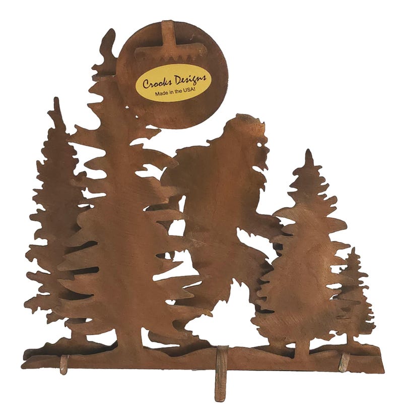 May include: Rustic metal wall decor featuring a silhouette of Bigfoot walking through a forest. The piece is made by Crooks Designs and is made in the USA.