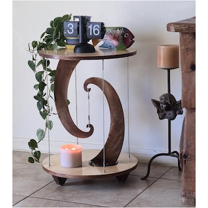 May include: A round two-tier side table featuring a light-colored wooden top and base connected by two curved, dark-toned wooden supports. The structure is held together by thin metal tension wires, creating a floating appearance for the top shelf.