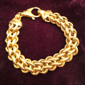 May include: A gold-colored bracelet with a double-strand chain design. The bracelet features a textured surface and a secure clasp. The jewelry is displayed against a dark burgundy background.