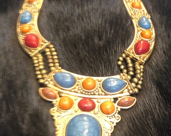 May include: Gold-toned statement necklace with colorful gemstone accents. The necklace features a large teardrop-shaped blue stone at the center, surrounded by smaller blue, orange, and red stones. The design is ornate and eye-catching, perfect for adding a pop of color to any outfit.