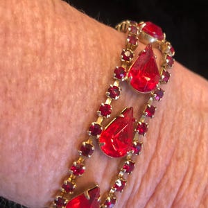 May include: Gold-toned bracelet with two rows of small, red rhinestones and three large, teardrop-shaped red gems. The bracelet is a vintage style, likely costume jewelry.