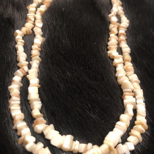 May include: A layered necklace made of small, irregularly shaped, off-white and tan shell pieces. The necklace is displayed against a dark, textured background, showcasing its natural color and texture. The necklace appears to be handmade.