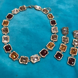 May include: A gold-toned necklace and matching earrings set. The jewelry features square-cut, multi-colored gemstones in shades of red, amber, clear, and brown. The necklace and earrings are displayed on a turquoise surface.