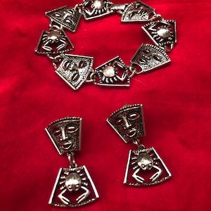 May include: Silver-toned jewelry set featuring a bracelet and a pair of earrings. The bracelet and earrings have a matching design with a mask-like face and a crab-like shape. The jewelry is displayed on a red background.
