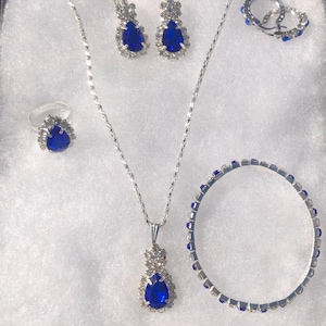 May include: A jewelry set featuring a silver necklace with a teardrop pendant, matching earrings, a ring, and a bracelet. Each piece is adorned with a large, vibrant blue gemstone surrounded by smaller clear stones. The set is displayed on a white, textured surface.