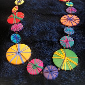 May include: A necklace featuring a series of circular wooden discs in various sizes and colors, including red, orange, yellow, green, blue, and purple. Each disc is wrapped with contrasting colored thread in a radial pattern and linked together.