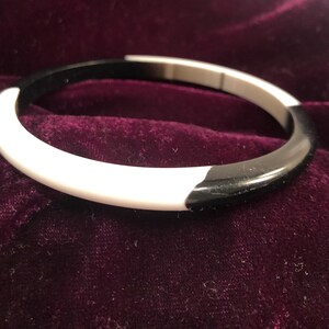 May include: A two-tone bangle bracelet. The bracelet is a circular shape, with a section of white and a section of black. The bracelet is made of a smooth, glossy material. The bracelet is resting on a dark purple velvet surface.