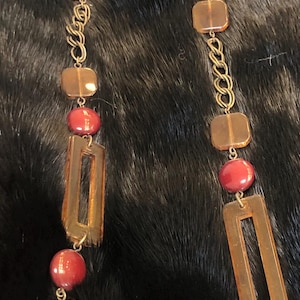 May include: A long necklace featuring alternating elements of rectangular amber-colored pendants, round maroon beads, and square amber-colored beads. The necklace is connected by gold-colored chain links. The jewelry is displayed against a dark, textured background.