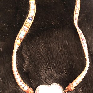 May include: A handmade necklace featuring a heart-shaped pendant with a floral design. The necklace has a braided leather cord adorned with colorful beads. The heart pendant is approximately 1 inch wide.